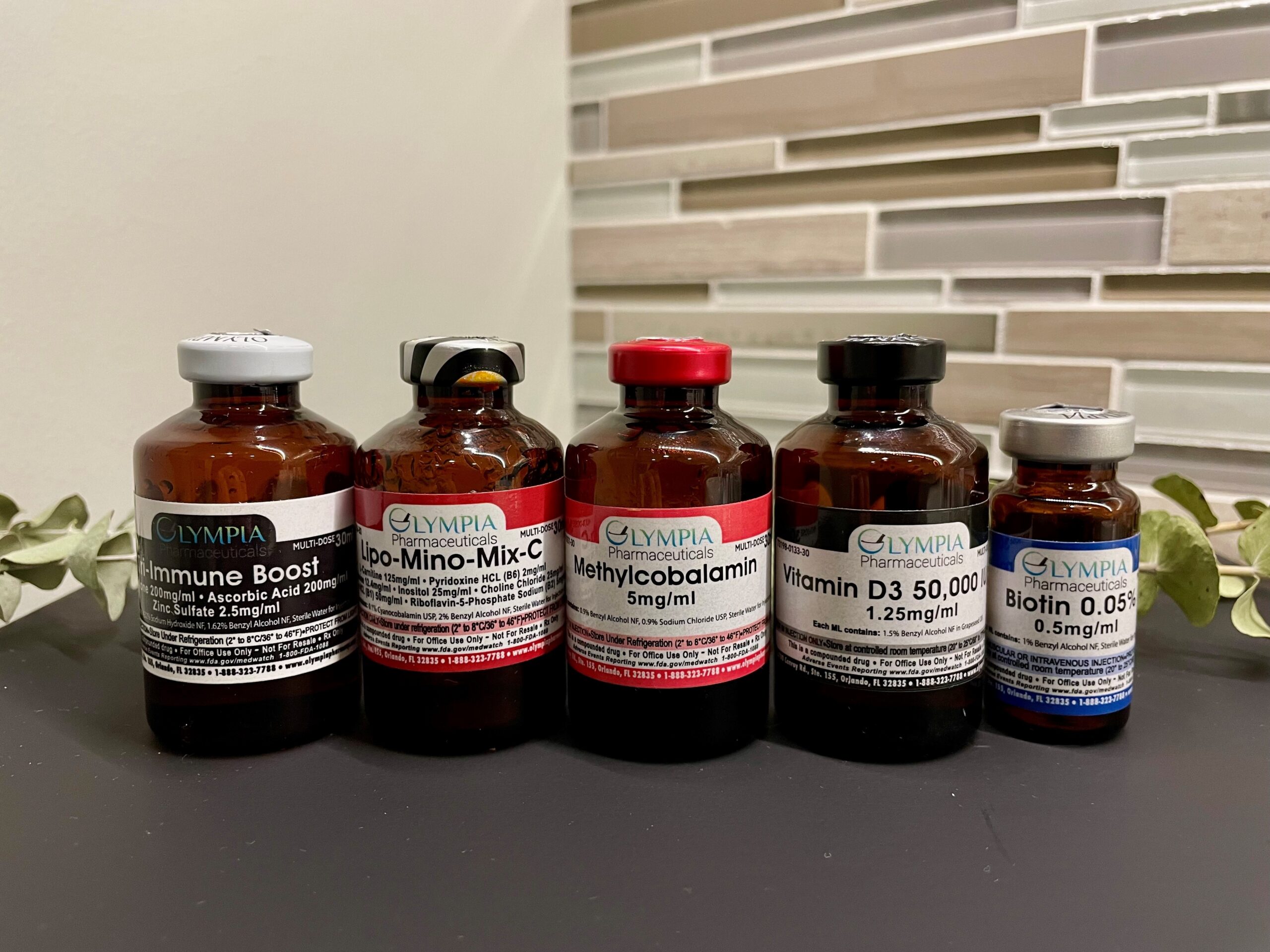 Five brown glass vials, reminiscent of those used for vitamin injection treatments, carry labels for various supplements, including Immune Boost, Lipo-Mino-Mix-C, Methylcobalamin, Vitamin D3 50,000 IU, and Biotin. - Anton's Salon and MSPA in Delafield, Wi